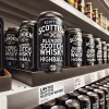 A Scottish blended Scotch whisky highball can with a black label displayed on shelves in a supermarket or convenience store. The setting shows its limited availability in a specific supermarket or convenience group. Simple store environment with visible branding, cans neatly aligned.